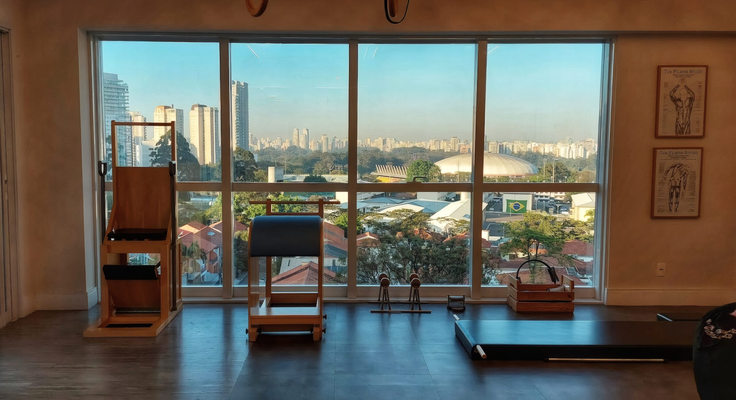Aparelhos clássicos de pilates diante da janela panorâmica do estúdio, com o horizonte de São Paulo ao fundo