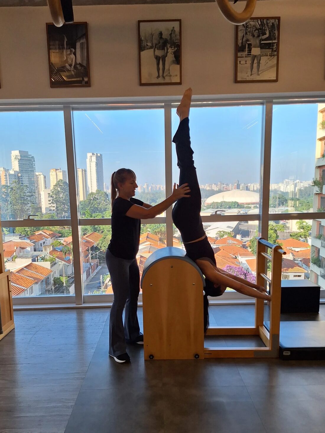 Professora guiando uma aluna em exercício invertido no Ladder Barrel, com São Paulo ao fundo pela janela